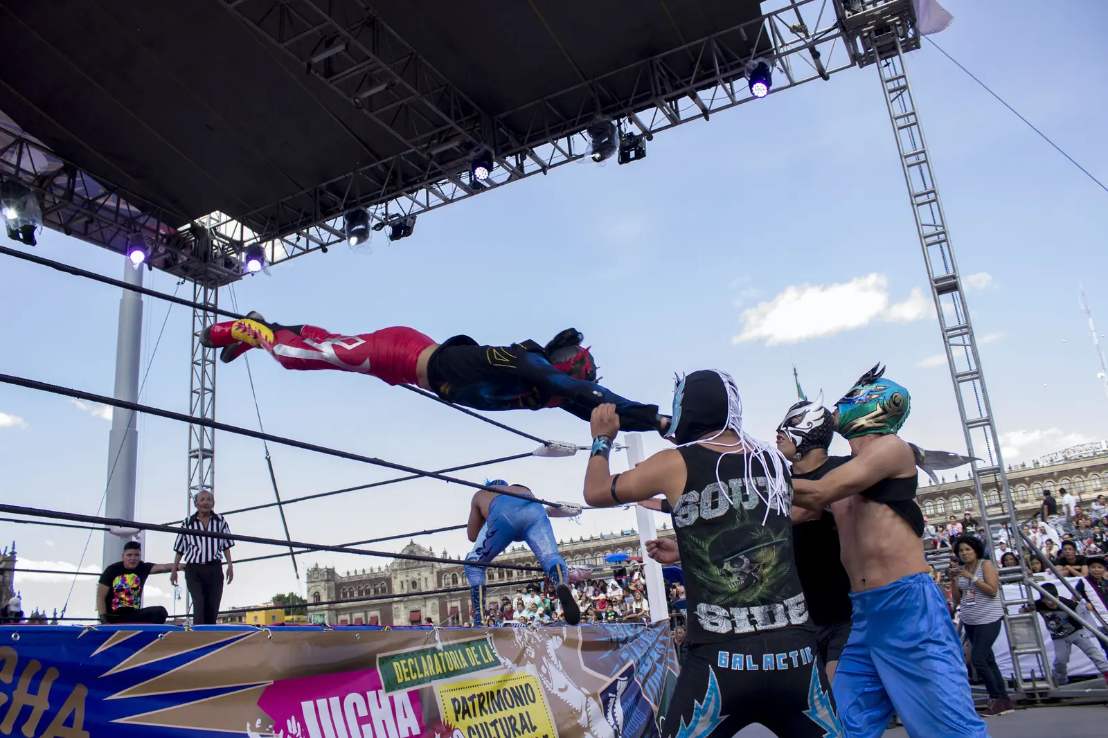 A luchador in red and black dives through the ropes of an open-air ring onto opponents as masked wrestlers watch beneath a blue sky