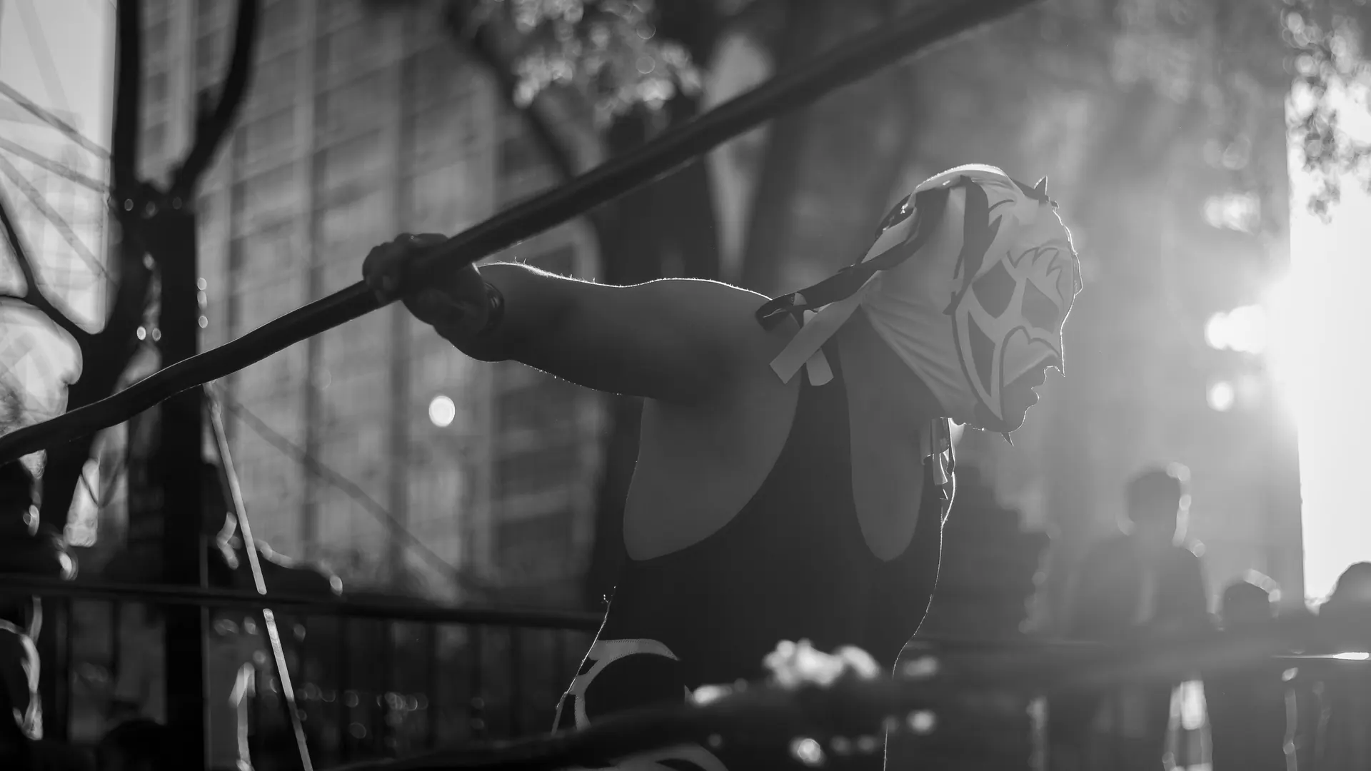 A masked luchador in a black singlet leans over the ropes of a ring, backlit by low sun, in a black and white photograph
