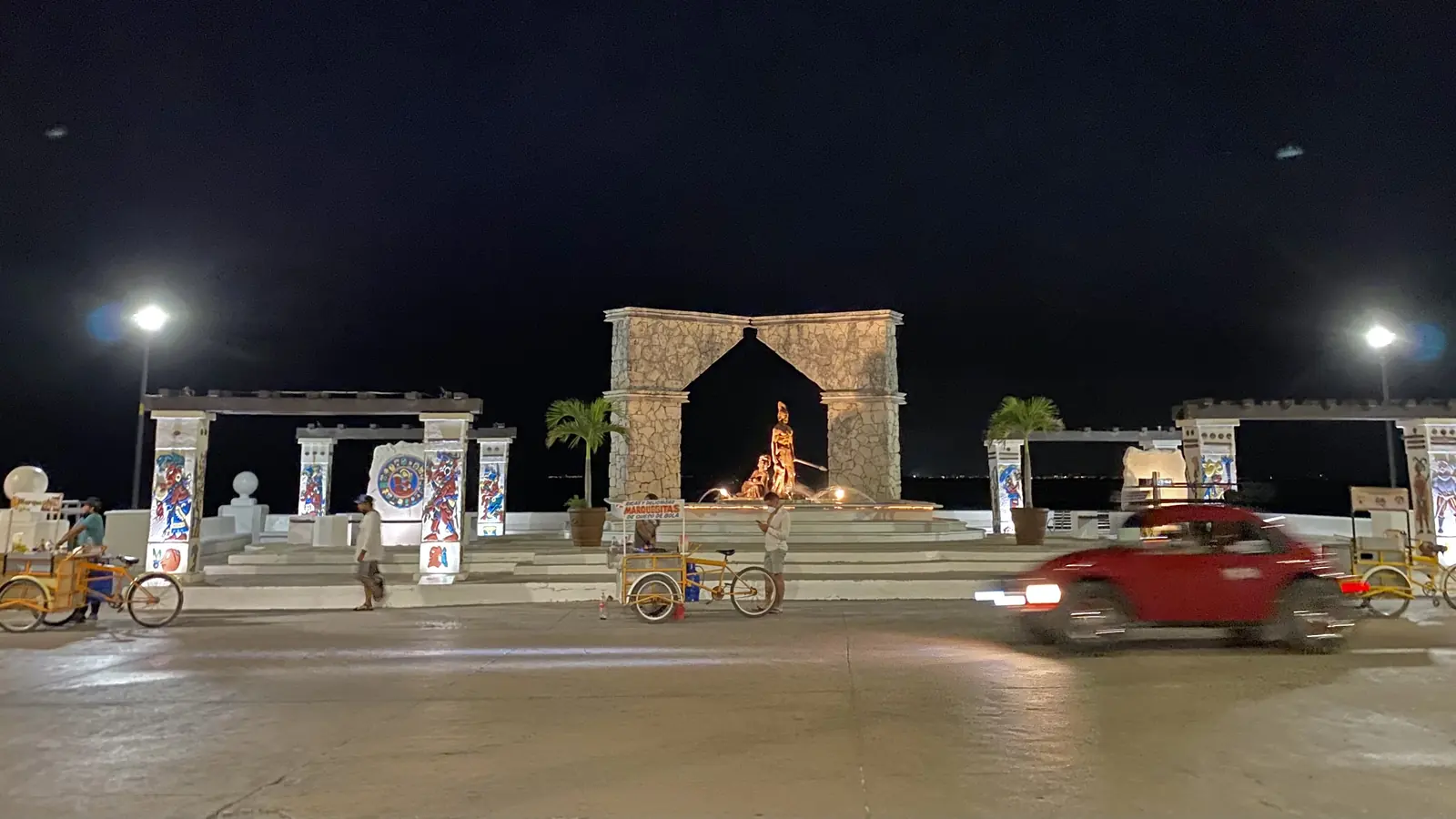 A stone arch and lit monuments on the San Miguel de Cozumel waterfront at night, with a red car passing and tricycle carts parked on the plaza