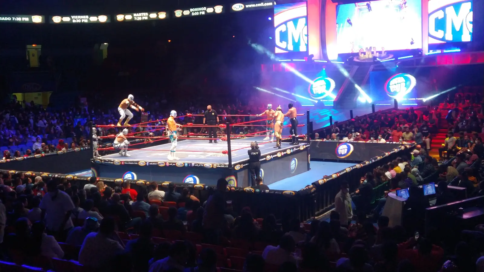 A lit lucha libre ring in a large arena, with luchadores in the ring, a referee and big video screens above